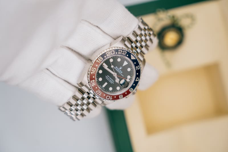 Close-up of a Rolex GMT-Master II “Pepsi” watch with a red and blue bezel and Jubilee bracelet, held in a white glove, with a green Rolex box blurred in the background.